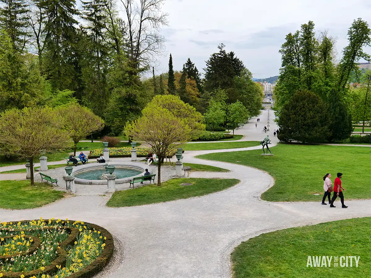 Tivoli Park in Ljubljana with winding gravel paths, green lawns, fountain and benches, people relaxing, and tree-lined promenade stretching into the distance.
