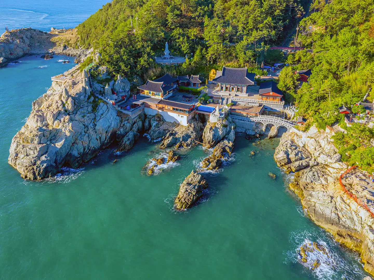 Aerial view of Haedong Yonggungsa Temple in Busan, perched on rocky coastline beside turquoise sea and surrounded by forested cliffs.