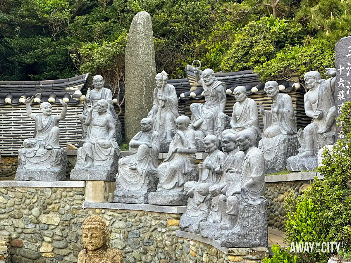 Stone statues of Buddhist disciples arranged around Haedong Yonggungsa Temple in Busan, with traditional walls and greenery surrounding the courtyard.