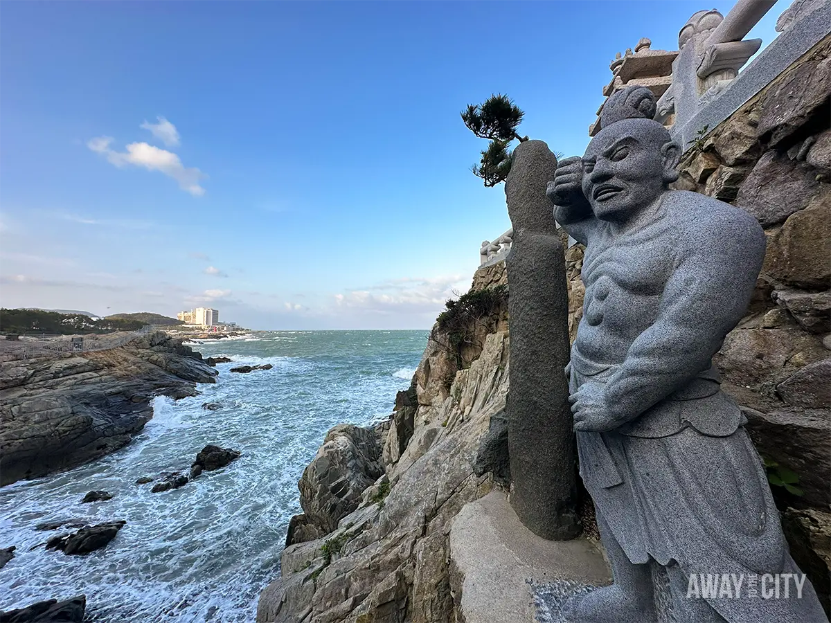 Stone guardian statue overlooking crashing waves and rocky coastline at Haedong Yonggungsa Temple in Busan under bright blue skies.