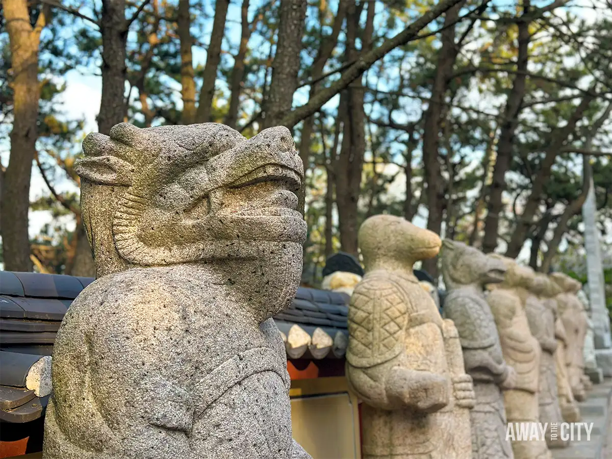 Close-up of carved zodiac animal statues lining pathway at Haedong Yonggungsa Temple in Busan, surrounded by pine trees and stone walls.