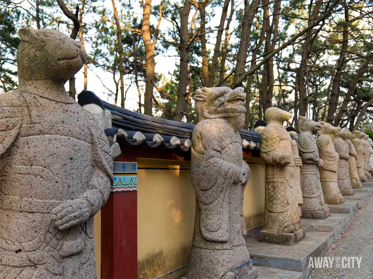 Row of carved zodiac animal statues lining pathway at Haedong Yonggungsa Temple in Busan beneath tall pine trees.