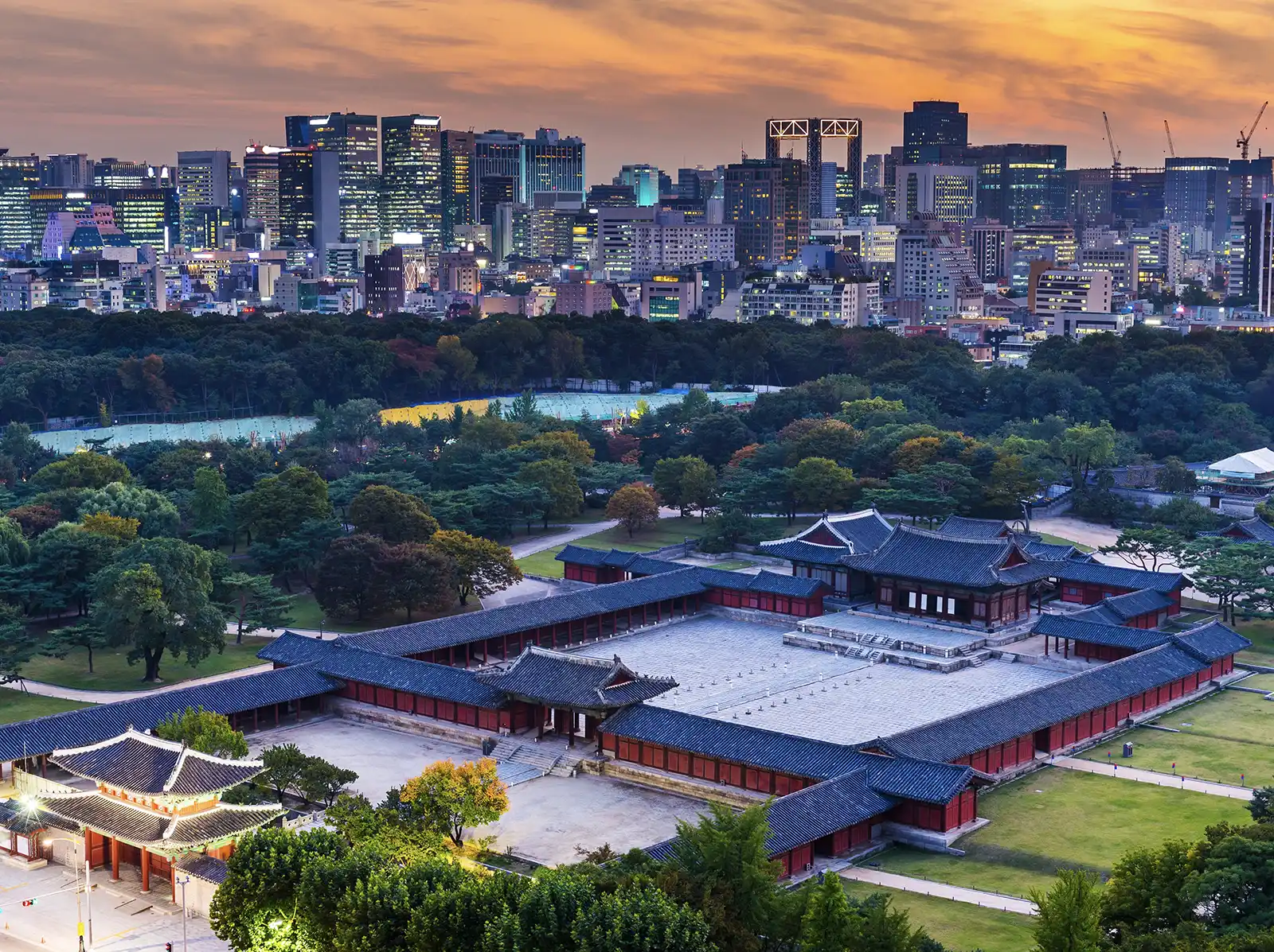 An aerial view of Changdeokgung Palace in Seoul, with the traditional red-roofed buildings set against a modern cityscape and sunset sky.