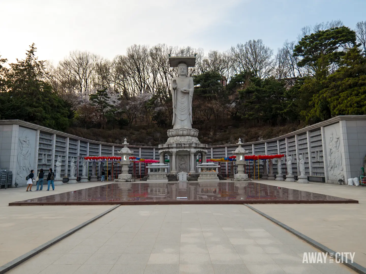 Tall stone Buddha statue standing in a memorial courtyard at Bongeunsa Temple in Seoul's Gangnam district, surrounded by lanterns, prayer walls and trees.