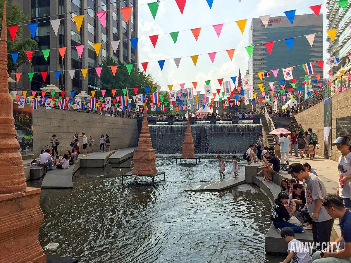 Visitors relaxing beside the Cheonggyecheon stream in Seoul, with colourful flags overhead, shallow water features and a cascading waterfall.