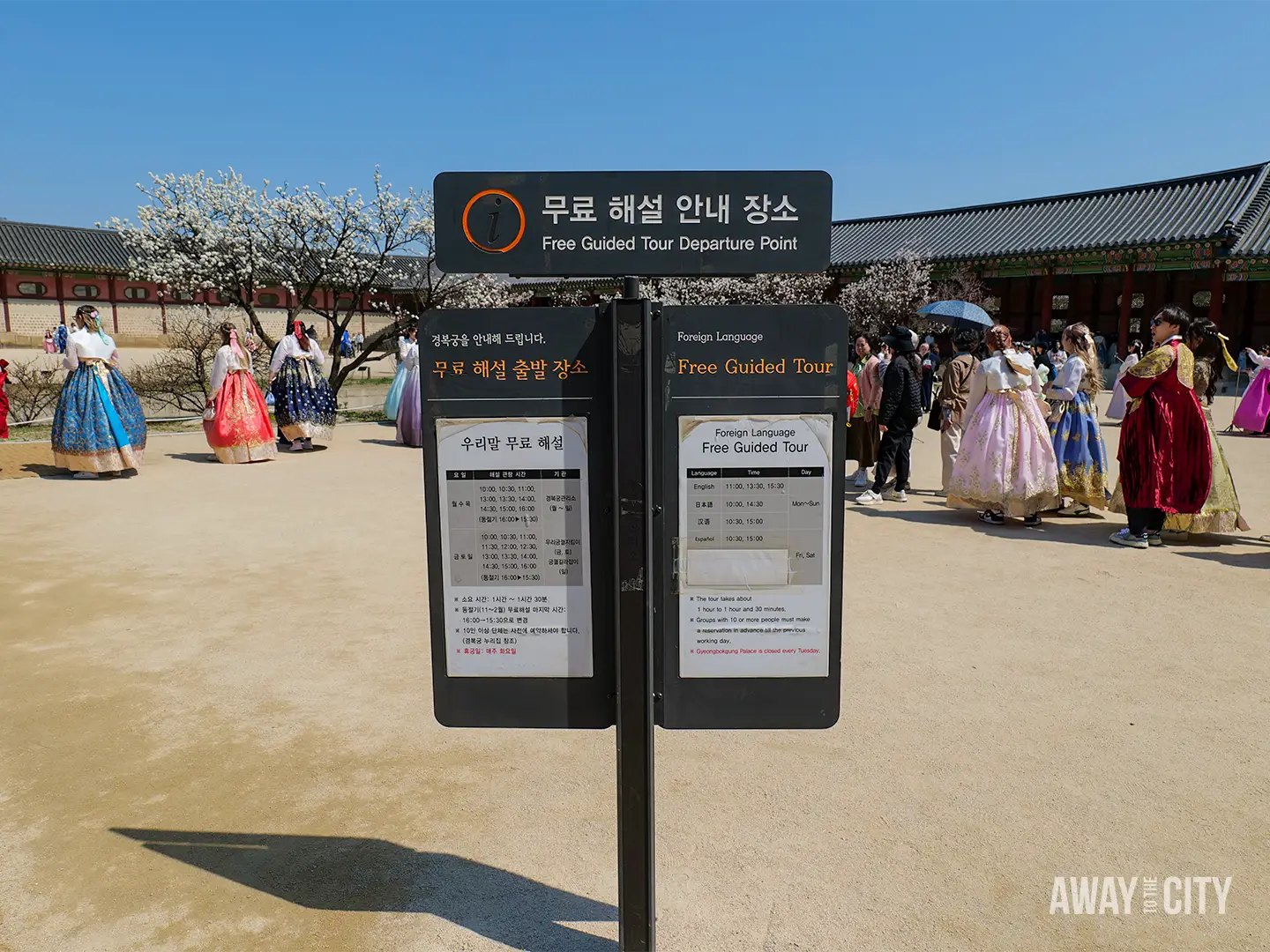 Sign reading “Free Guided Tour Departure Point” in Korean and English at Gyeongbokgung Palace, with visitors wearing colourful hanbok in the background.