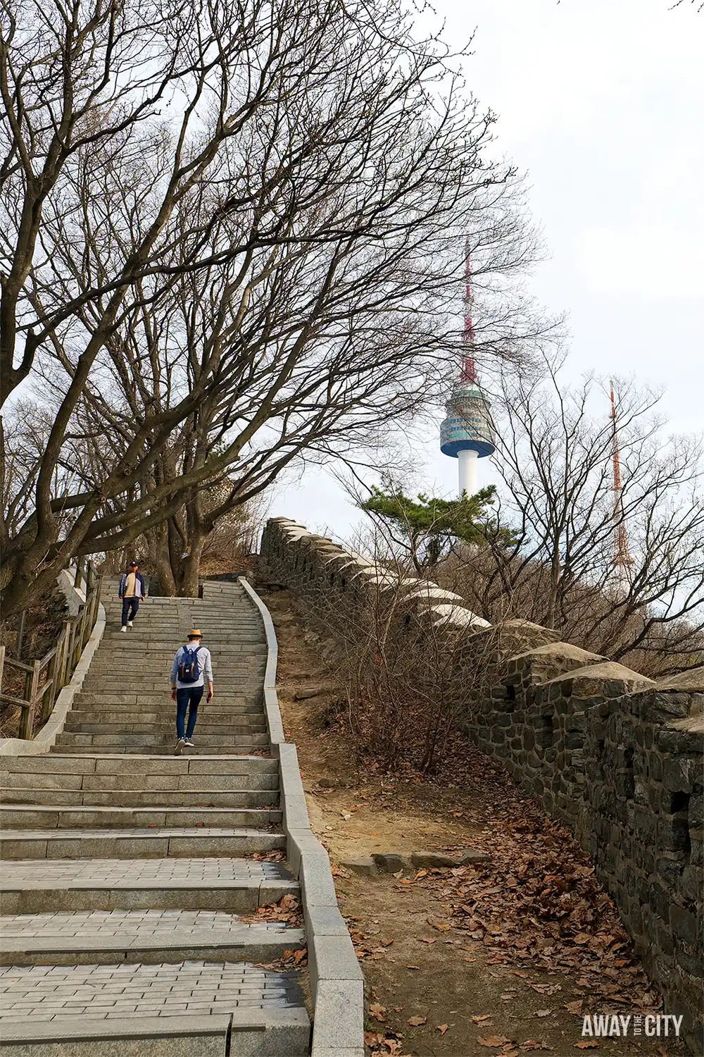 Stone staircase through Namsan Park leading towards N Seoul Tower, with historic walls, bare trees and hikers ascending.