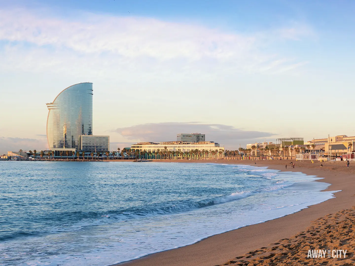 The sandy shore of Barceloneta Beach with the W Barcelona hotel and the Mediterranean Sea waves washing in.