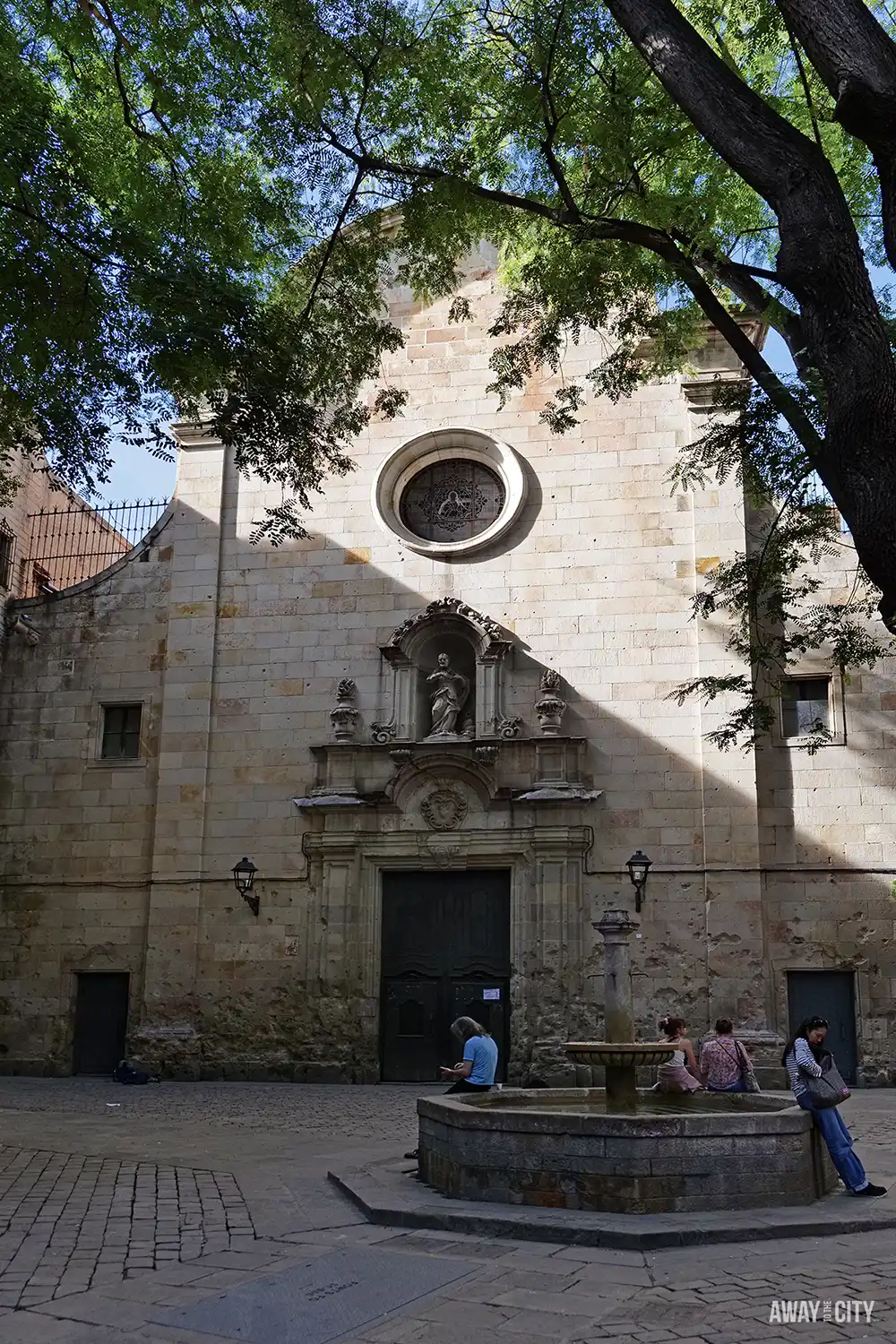 The baroque facade of the Church of Saint Philip Neri in Barcelona, framed by trees, with people sitting by a fountain.