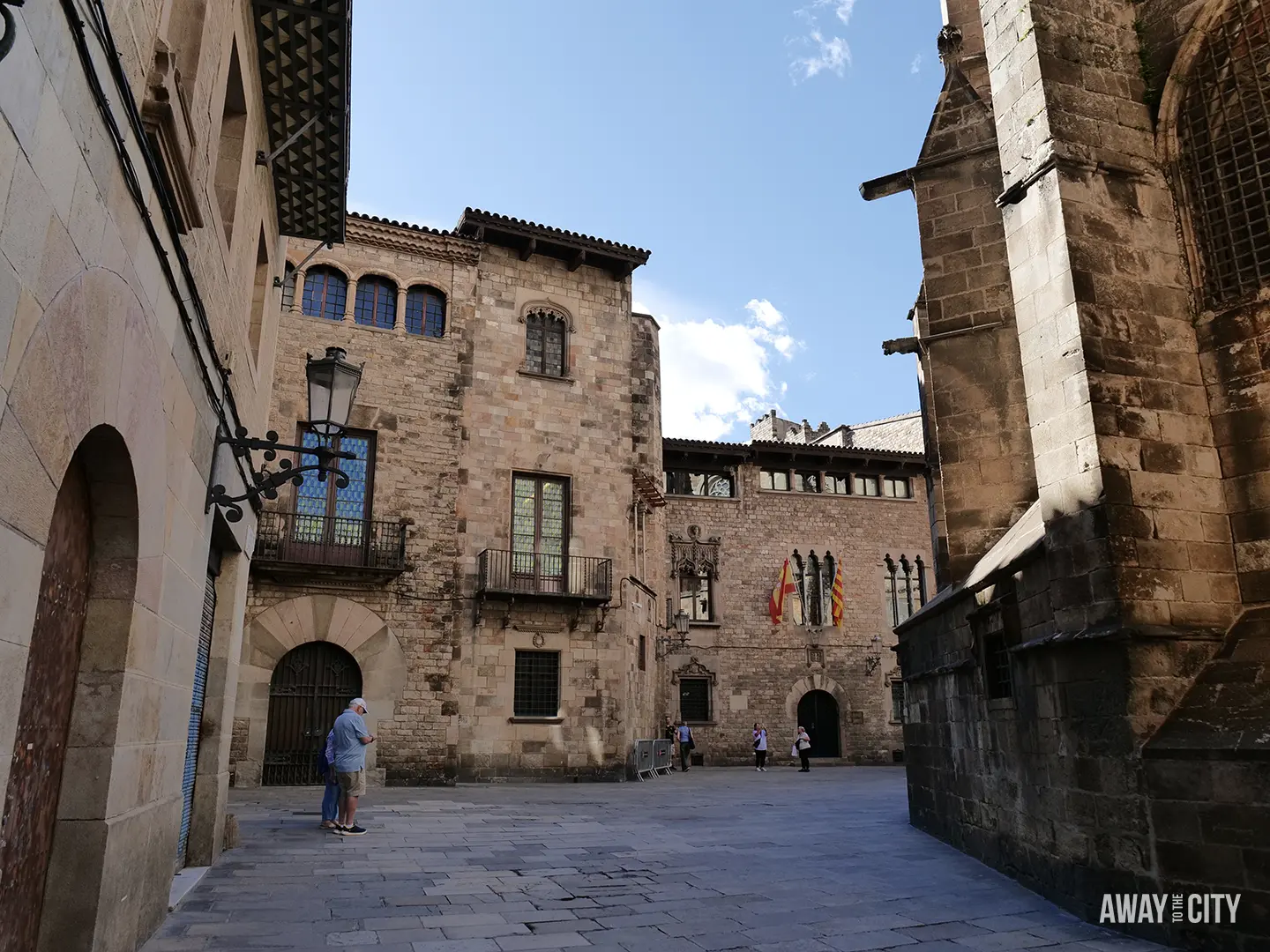 A sunlit, empty plaza in the Barri Gòtic of Barcelona, surrounded by medieval stone buildings and a few people.