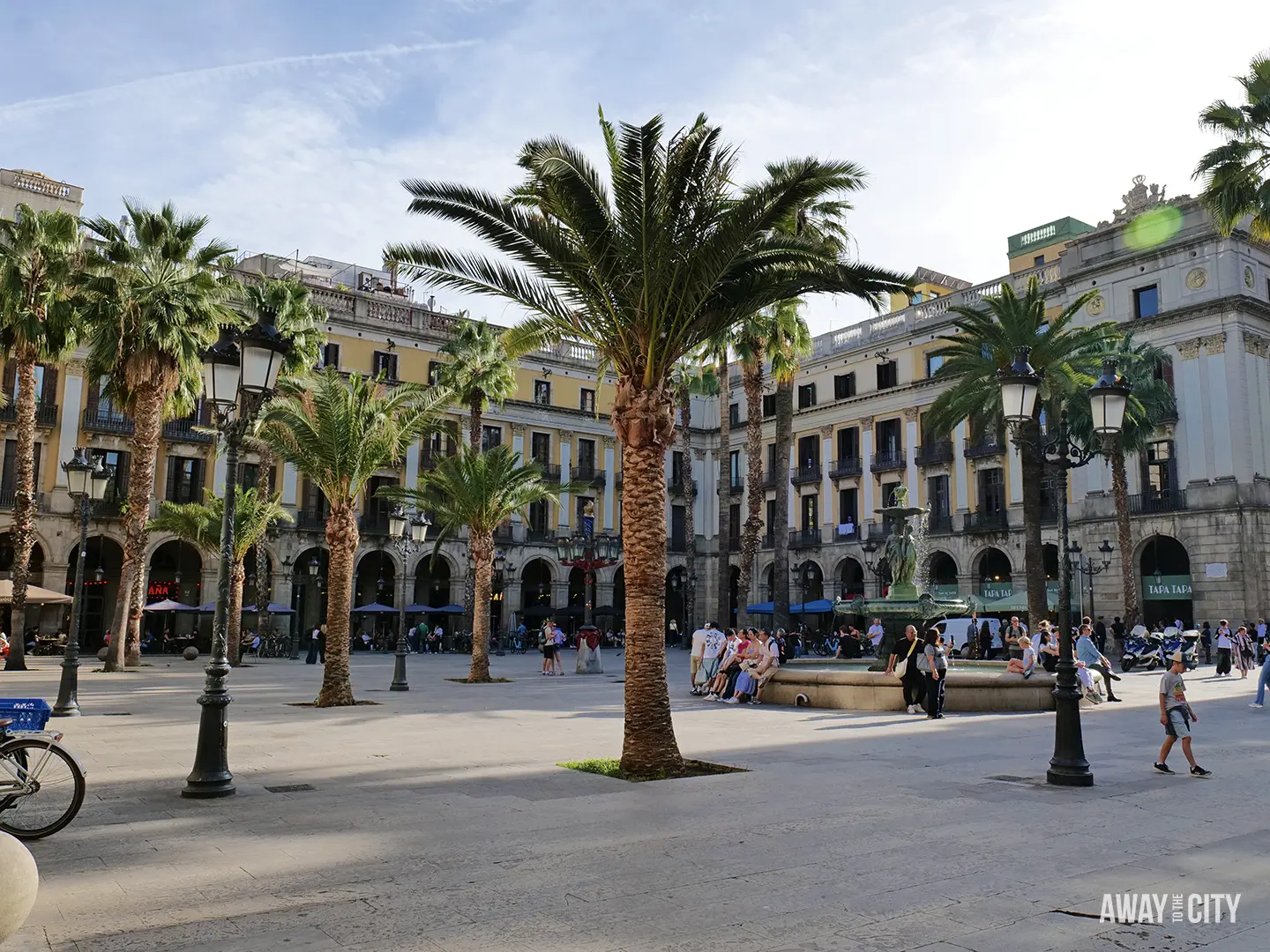 The sunny Plaça Reial in Barcelona, featuring a central fountain, streetlights, palm trees, and surrounding yellow buildings.