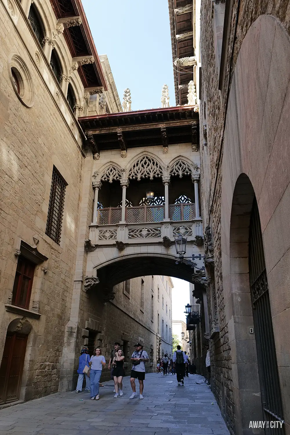 A few people walk beneath the Gothic Revival bridge connecting buildings in the Barri Gòtic district of Barcelona.