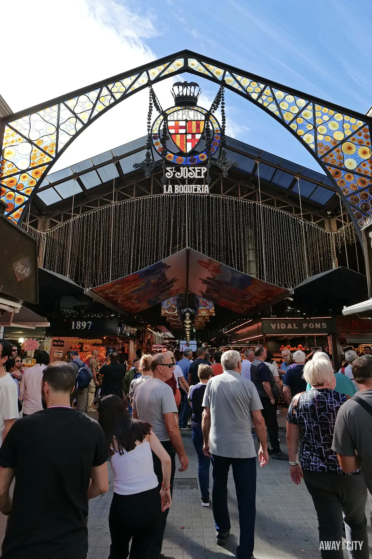 A crowd of people enters Barcelona's La Boqueria Market through the ornate, stained-glass iron archway under a blue sky.