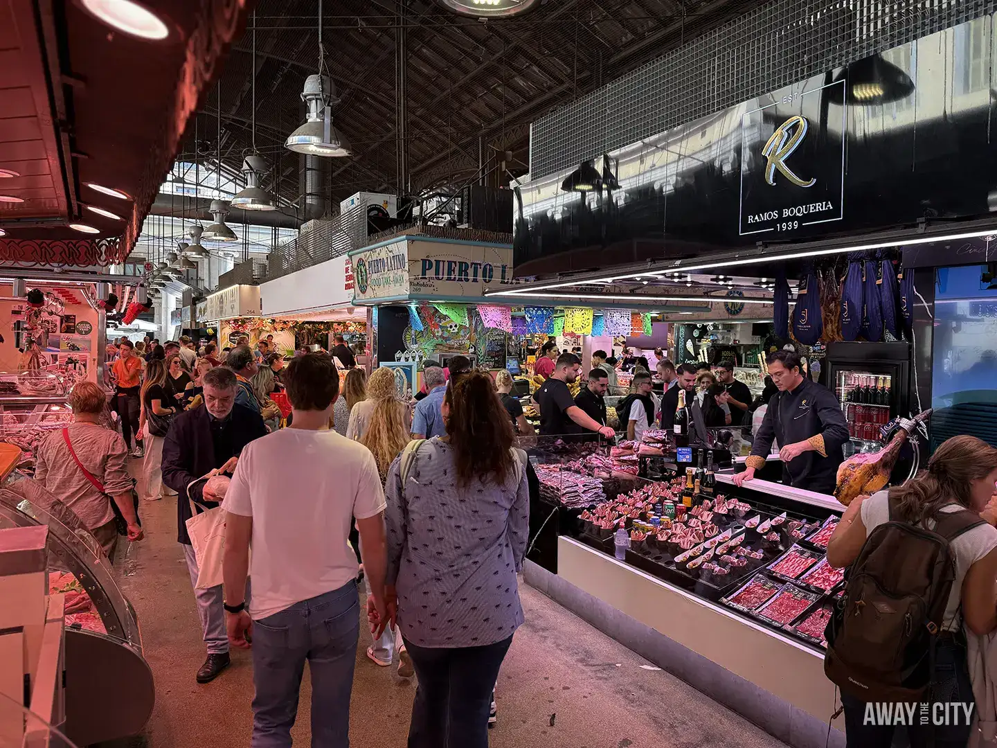 A bustling interior view of Mercat de la Boqueria in Barcelona, showing people walking past stalls selling meat and cured ham.