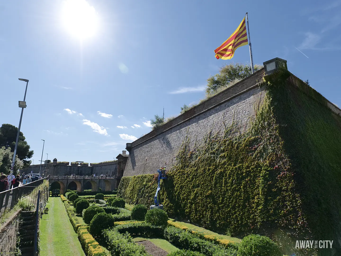 The exterior wall of Montjuïc Castle in Barcelona, covered in ivy, with the Catalan flag flying above formal gardens.