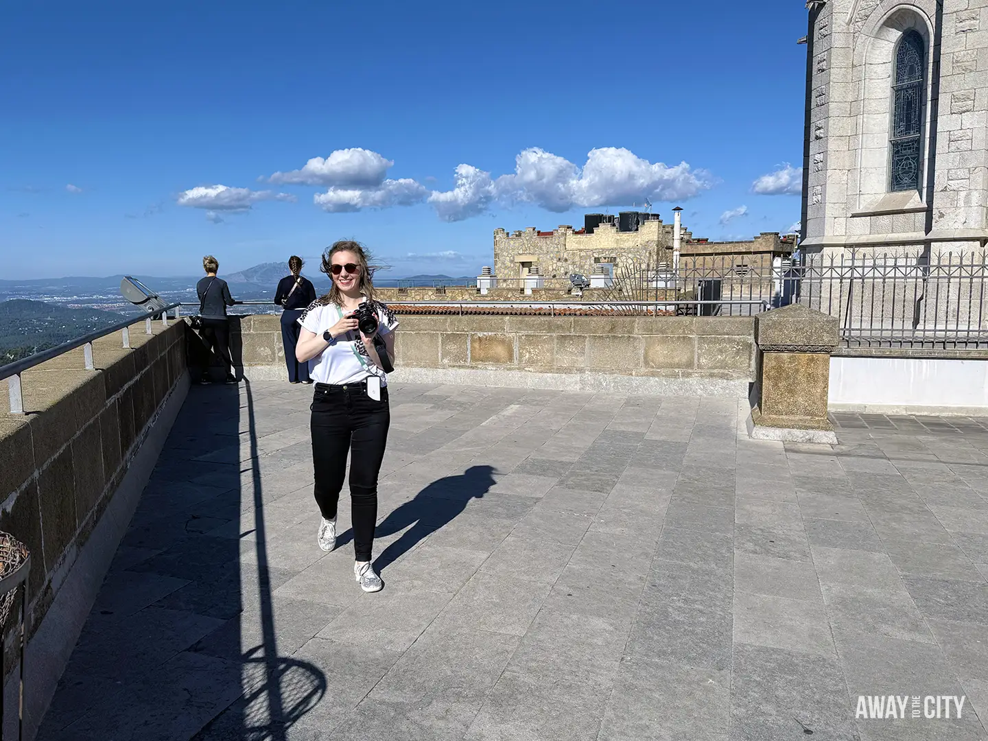 A smiling person stands on a sunny, stone viewing terrace near the Tibidabo Church in Barcelona, with a distant view of the city.
