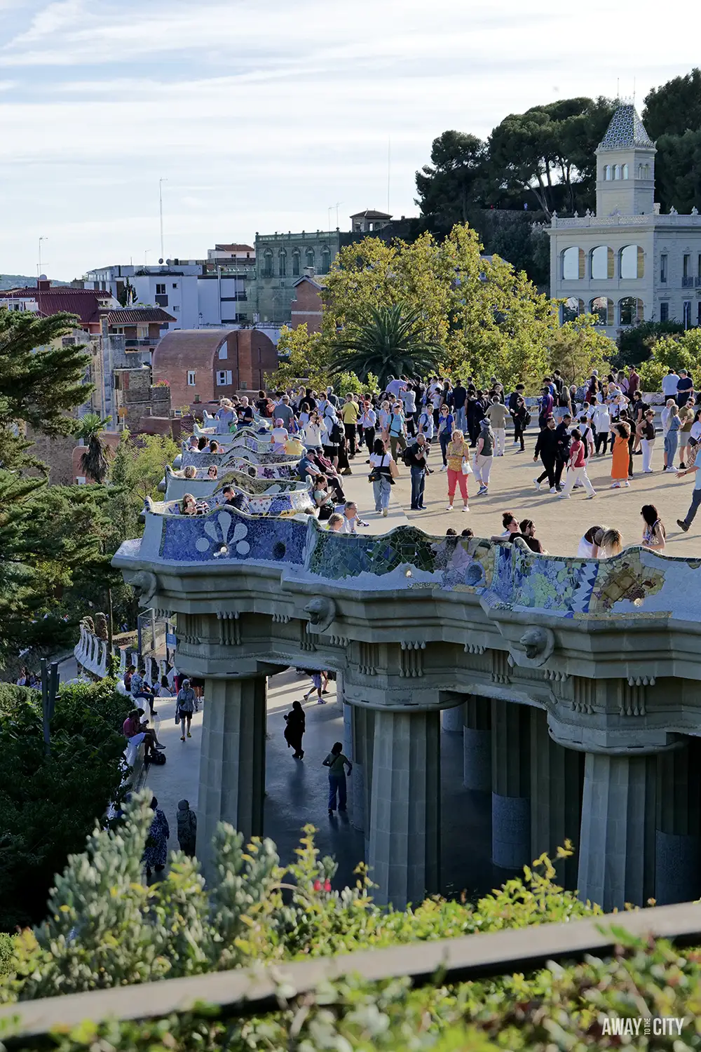 A sunny view of a crowded Park Güell terrace with a tiled serpentine bench overlooking the Hypostyle Room columns.