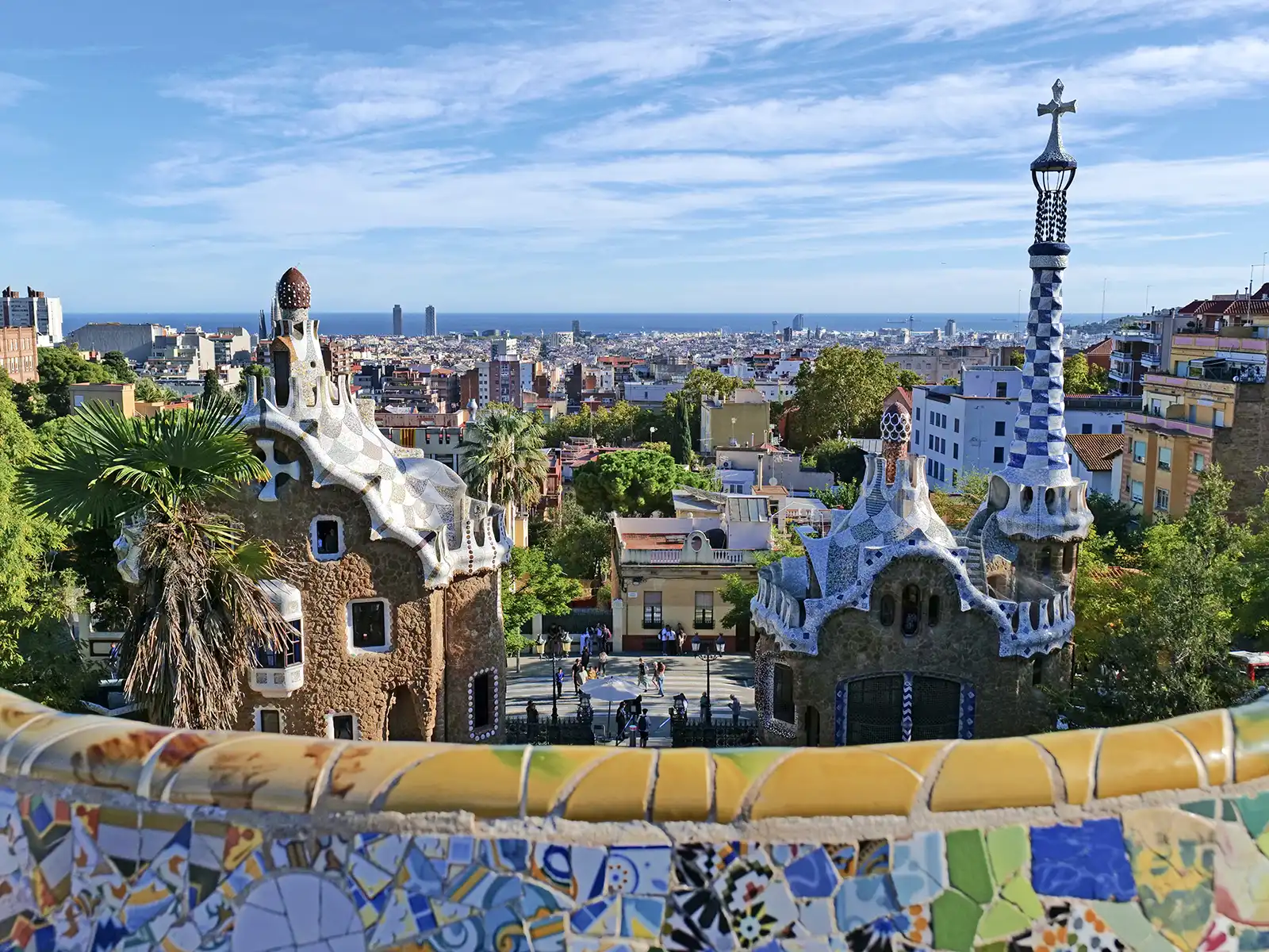 A sunny view from Park Güell shows two whimsical buildings and the city of Barcelona stretching to the sea.