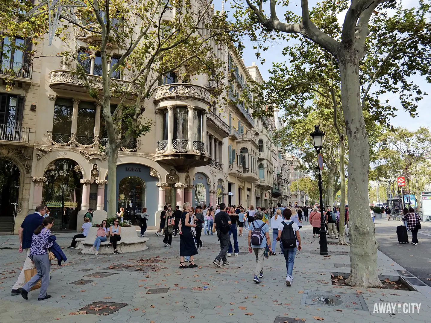 Shoppers and tourists walk along the Passeig de Gràcia in Barcelona, lined with plane trees and modernist buildings.
