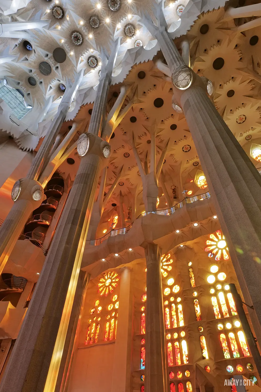 An angular view of the Sagrada Família interior shows the warm light from the stained glass on the towering, geometric columns.