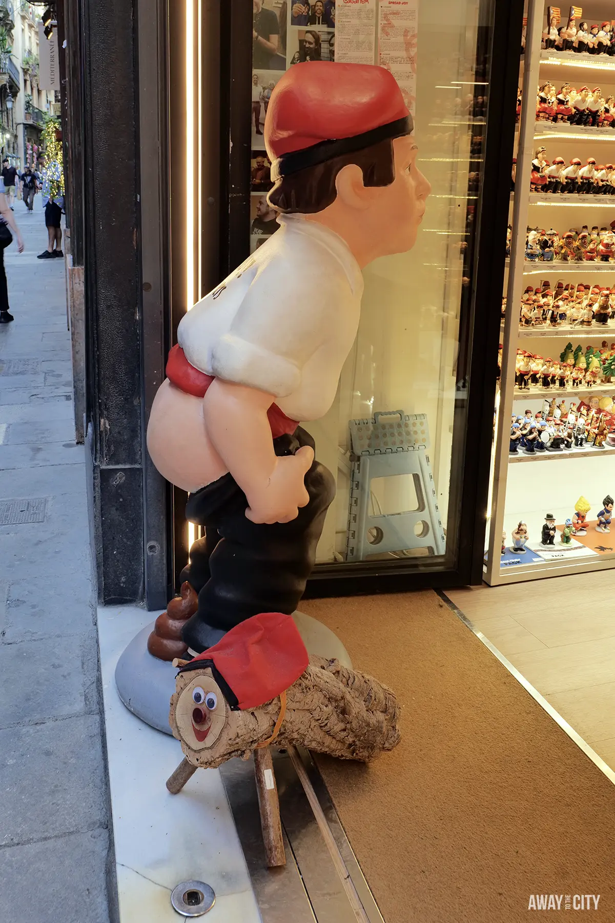 A large, comical display of a Caganer figurine, showing its trousers down, next to a Caga Tió log on a shop doorway.