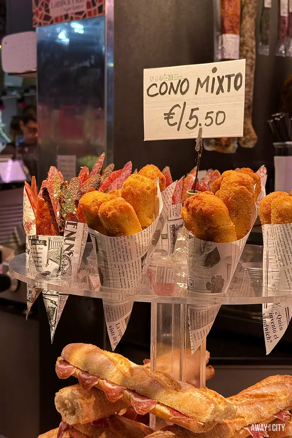 A market stall display shows paper cones of croquettes and sausages.