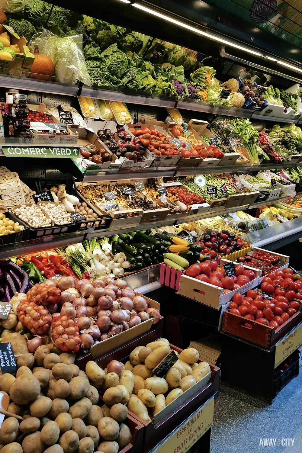 A well-stocked display in a greengrocer's shows boxes of fresh local produce, including potatoes, onions, and tomatoes.