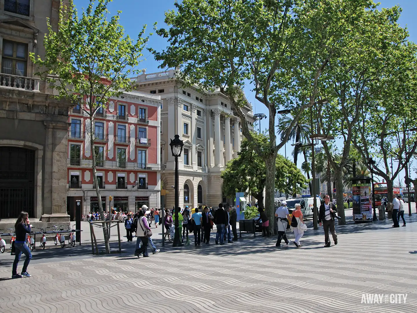A busy, sunny scene in Barcelona showing Plaça del Portal de la Pau with the Govern Militar building and people walking on the wavy-patterned paving.