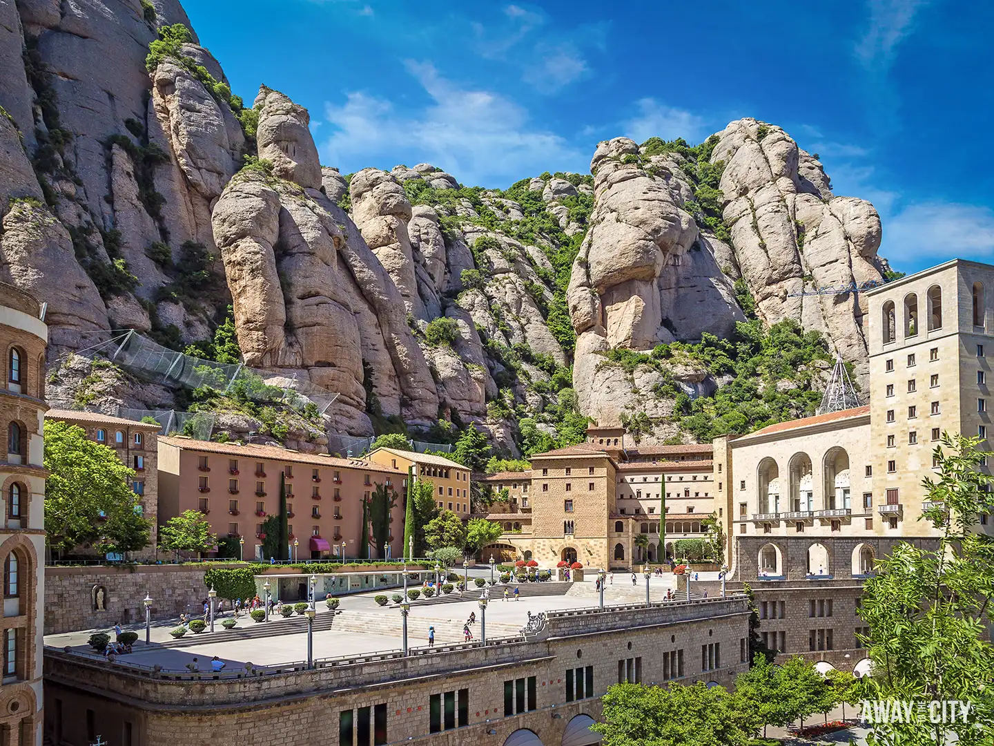 A bright, sunny view of the Santa Maria de Montserrat abbey nestled against the towering, sculpted mountains.