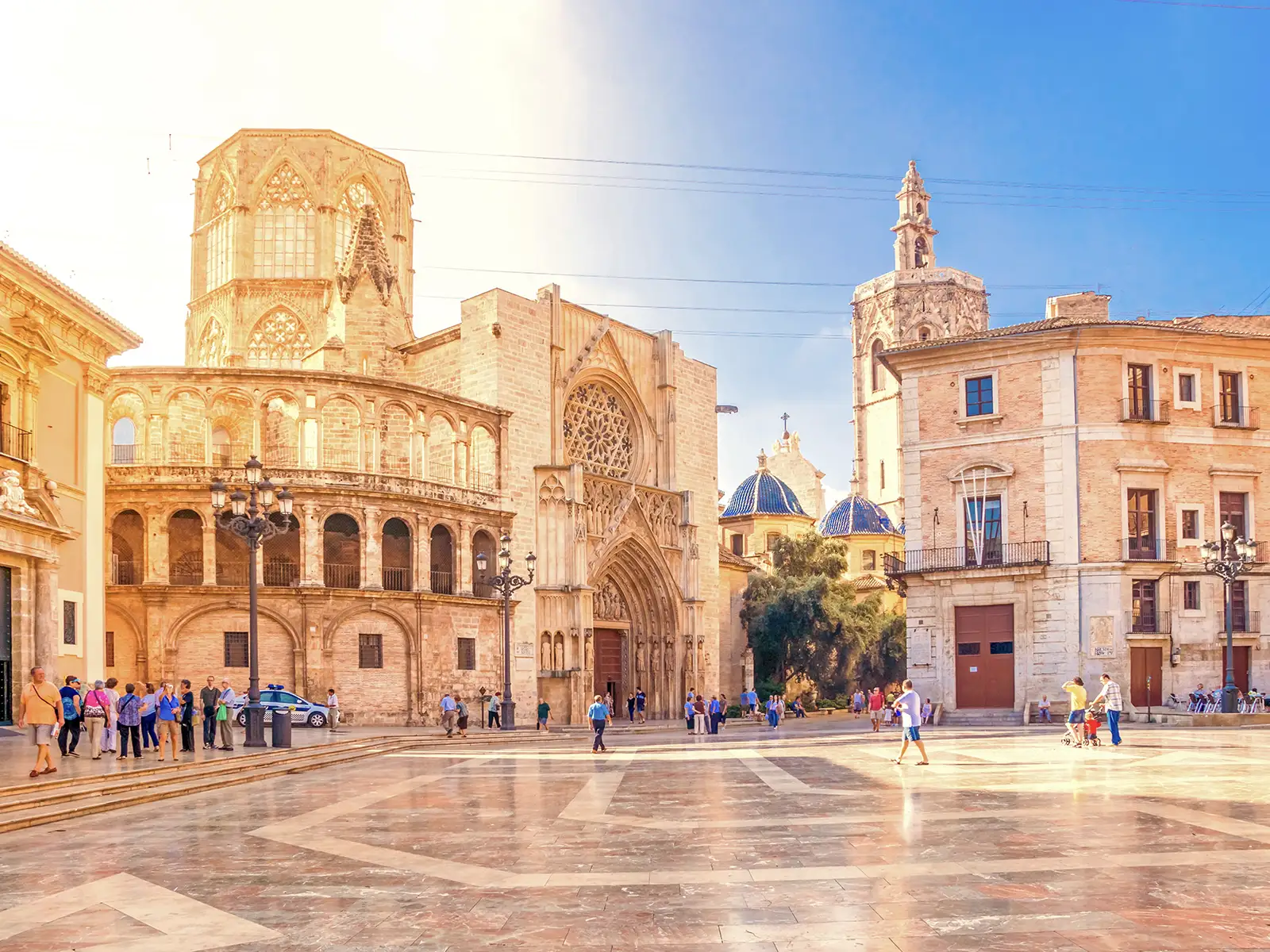 A sunlit square in Valencia with a group of tourists and the city's cathedral in the background.