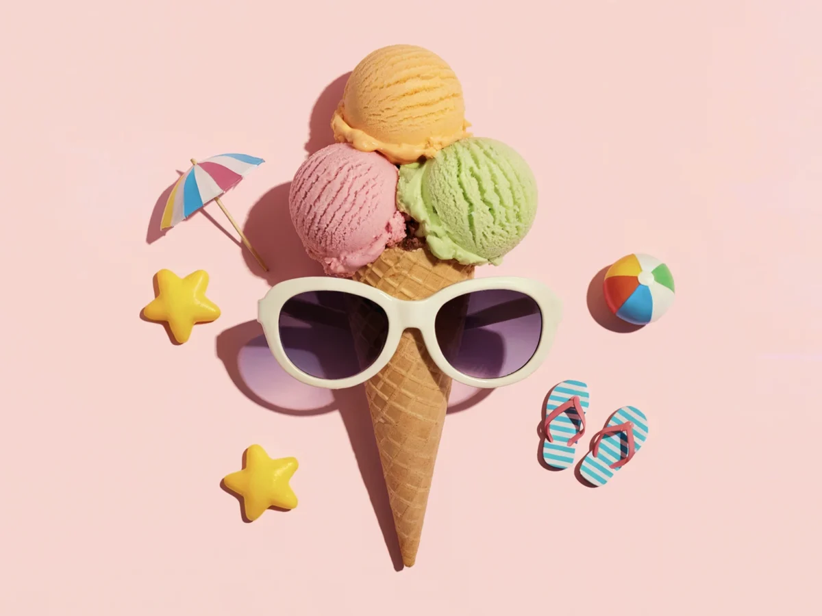 Flat lay of a colourful ice cream cone with sunglasses and mini summer props on a pastel pink background.