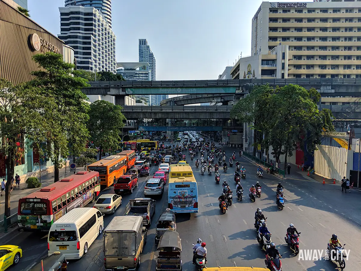 Busy Bangkok intersection with cars, buses and motorbikes beneath elevated walkways, surrounded by modern buildings and roadside trees.