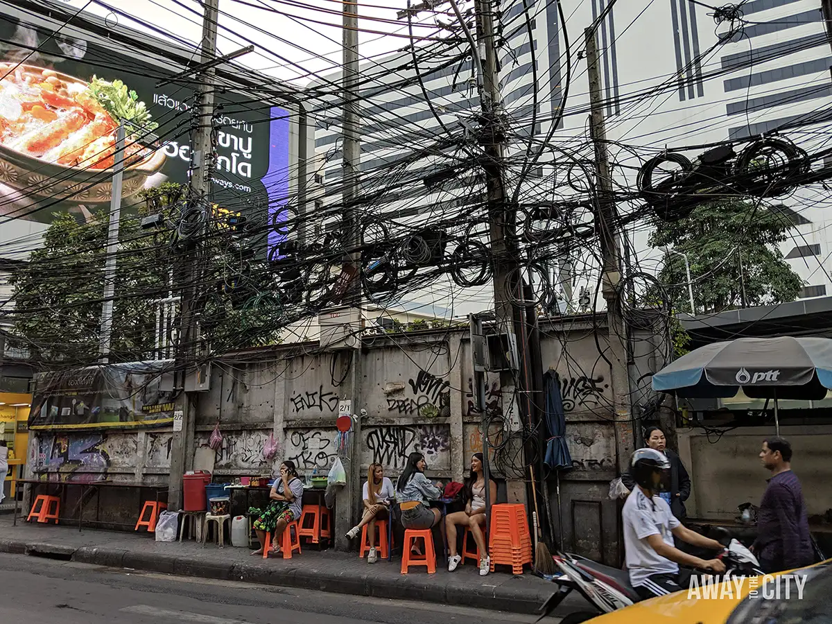 Street-side seating beneath dense overhead power cables, with people on plastic stools, passing motorbikes, graffiti walls and tall city buildings behind.