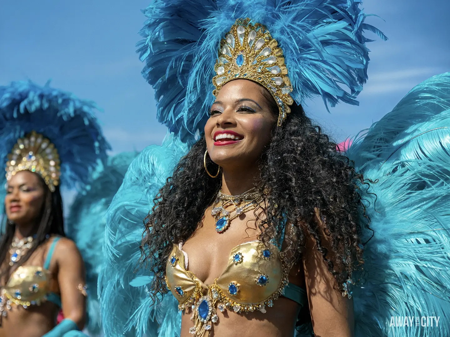 A woman in a vibrant blue feather headdress and a gold bikini top smiles, with another person in similar attire behind her, at the Toronto Caribbean Carnival.