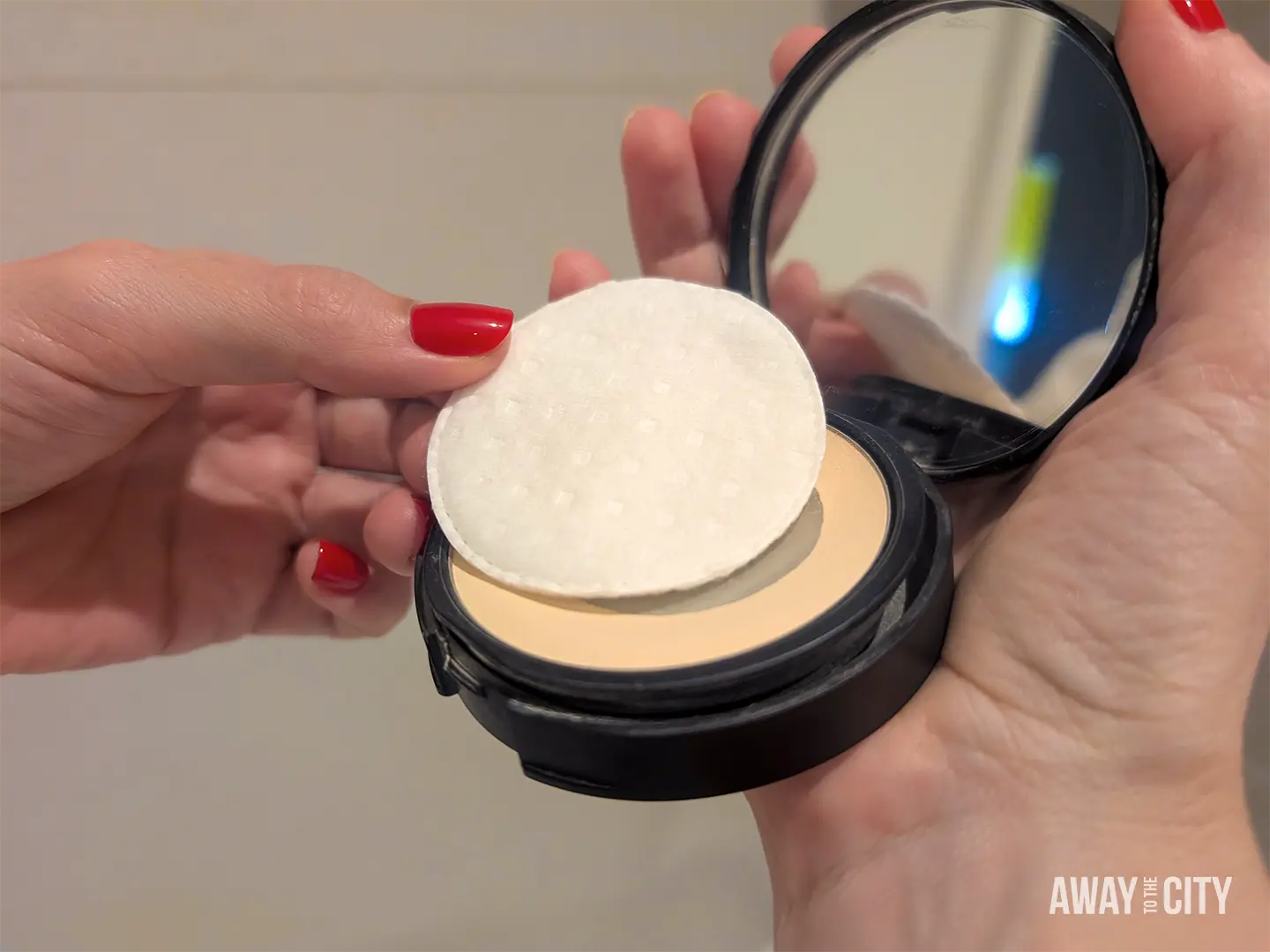 Hands holding an open powder compact with a cotton pad placed over the pressed powder, which is an easy packing technique to prevent breakage during travel.
