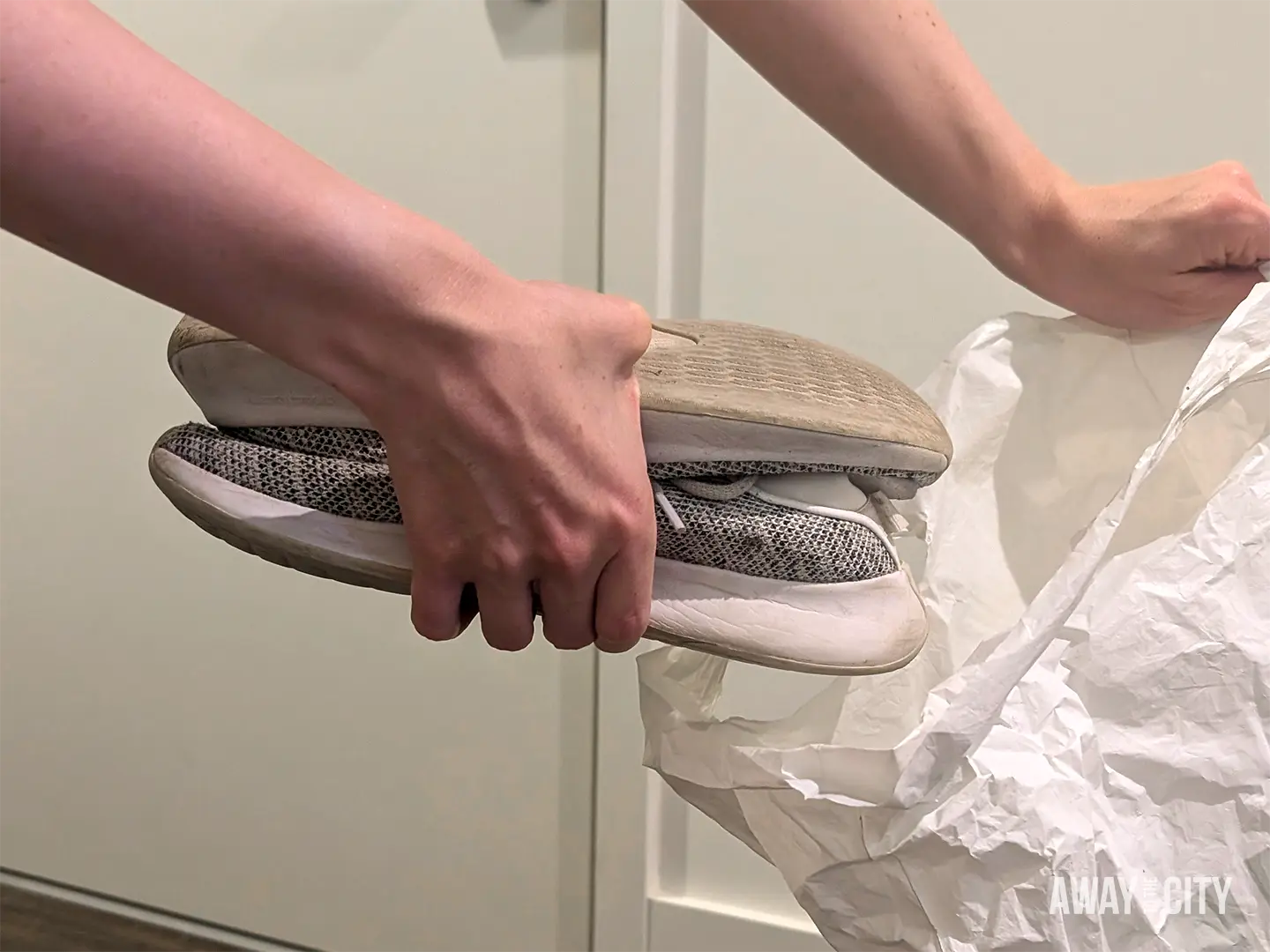 Hands placing a pair of beige trainers inside a plastic bag to protect other belongings when packing a suitcase.