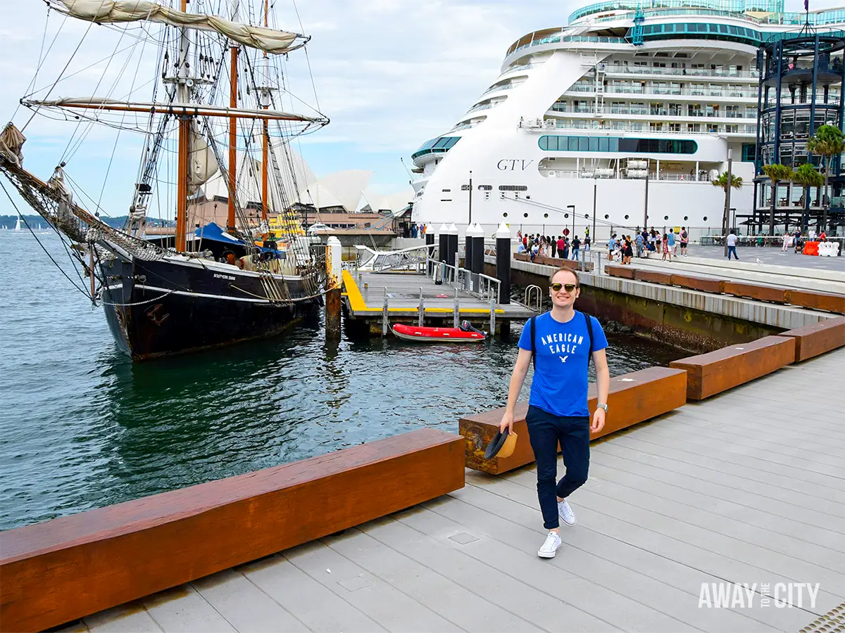 Person walking along Sydney Harbour waterfront beside a historic tall ship and large cruise ship docked near the city.