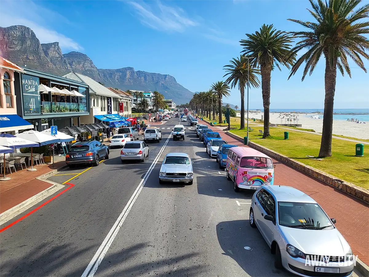 Victoria Road along Camps Bay beachfront in Cape Town, with palm trees, sandy beach and the distinctive Twelve Apostles mountain range rising behind.