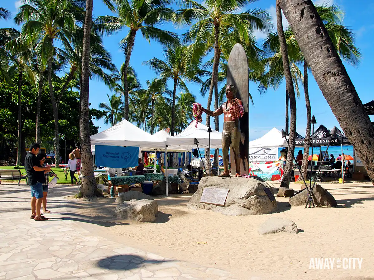 Statue of legendary Hawaiian surfer Duke Kahanamoku at Waikiki Beach in Honolulu, surrounded by palm trees and surfboards beneath a bright blue sky.