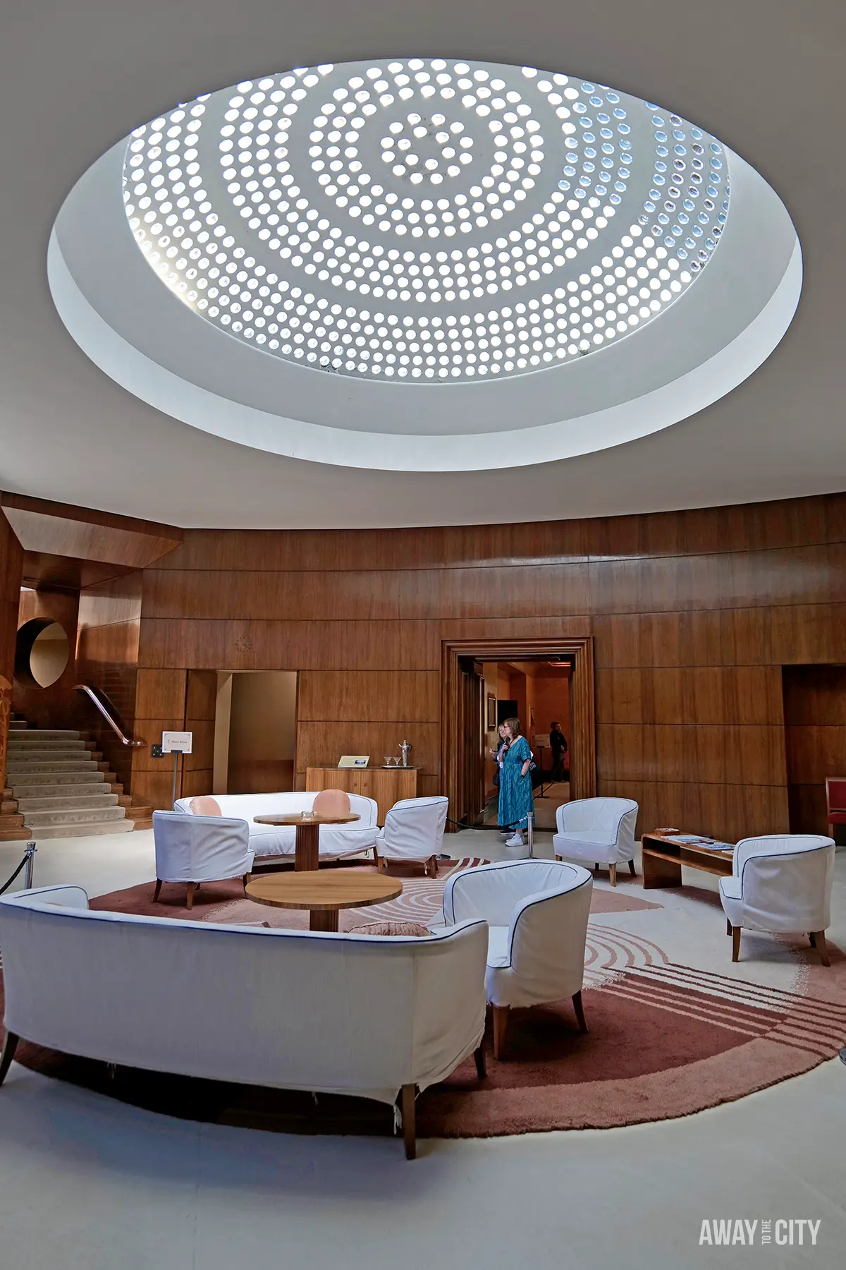 The Art Deco entrance hall of Eltham Palace features wood-panelled walls, white furniture, a circular patterned skylight, and a red circular rug, offering a glimpse into a potential 1-day trip from London by train.