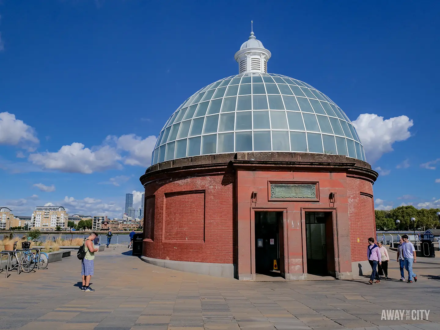 The round, red-brick entrance building to the Greenwich Foot Tunnel, with a glass dome roof against a blue sky.