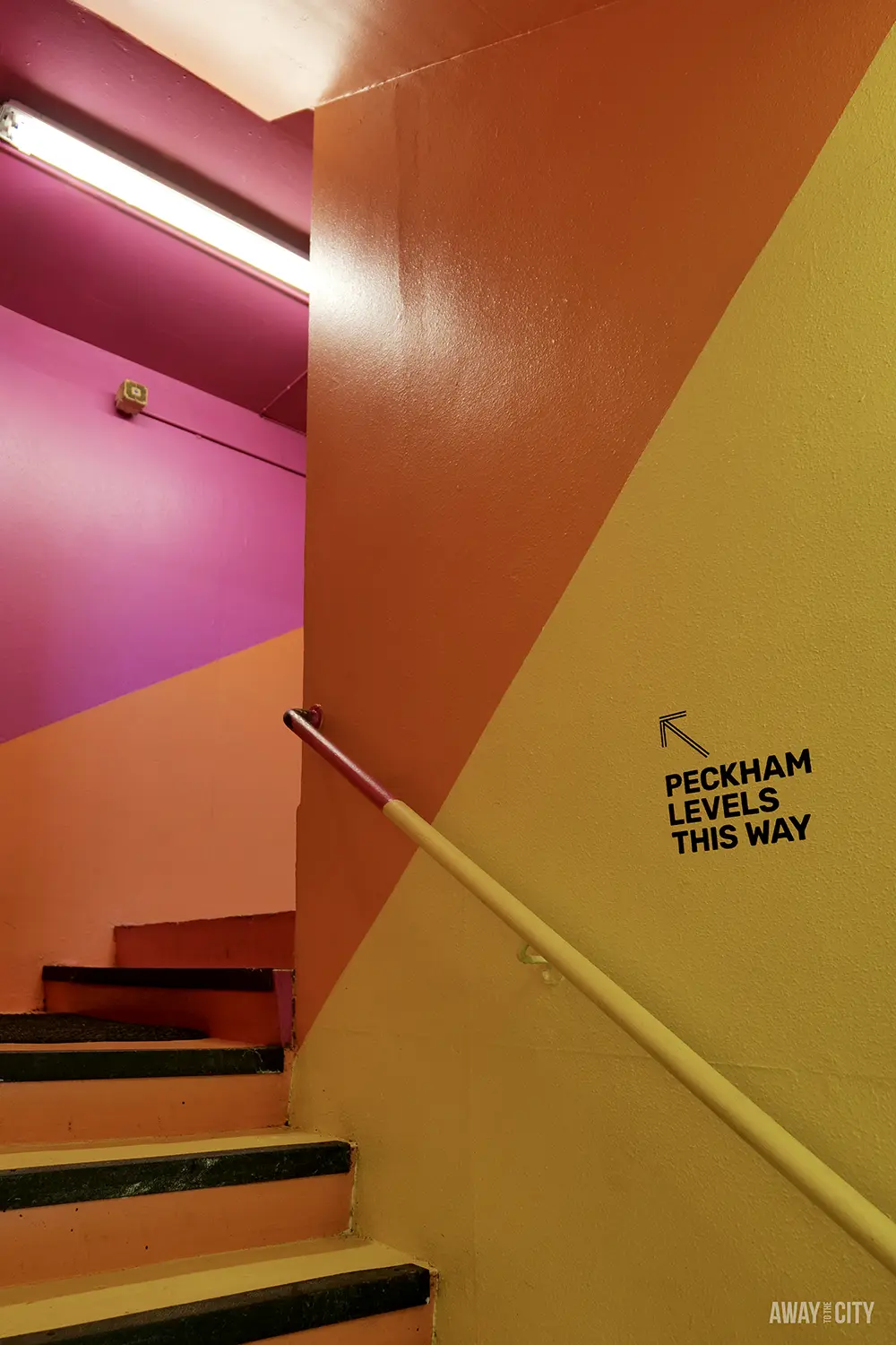 A colourful staircase painted with diagonal stripes of yellow, orange, and pink, with a sign pointing to "Peckham Levels this way".
