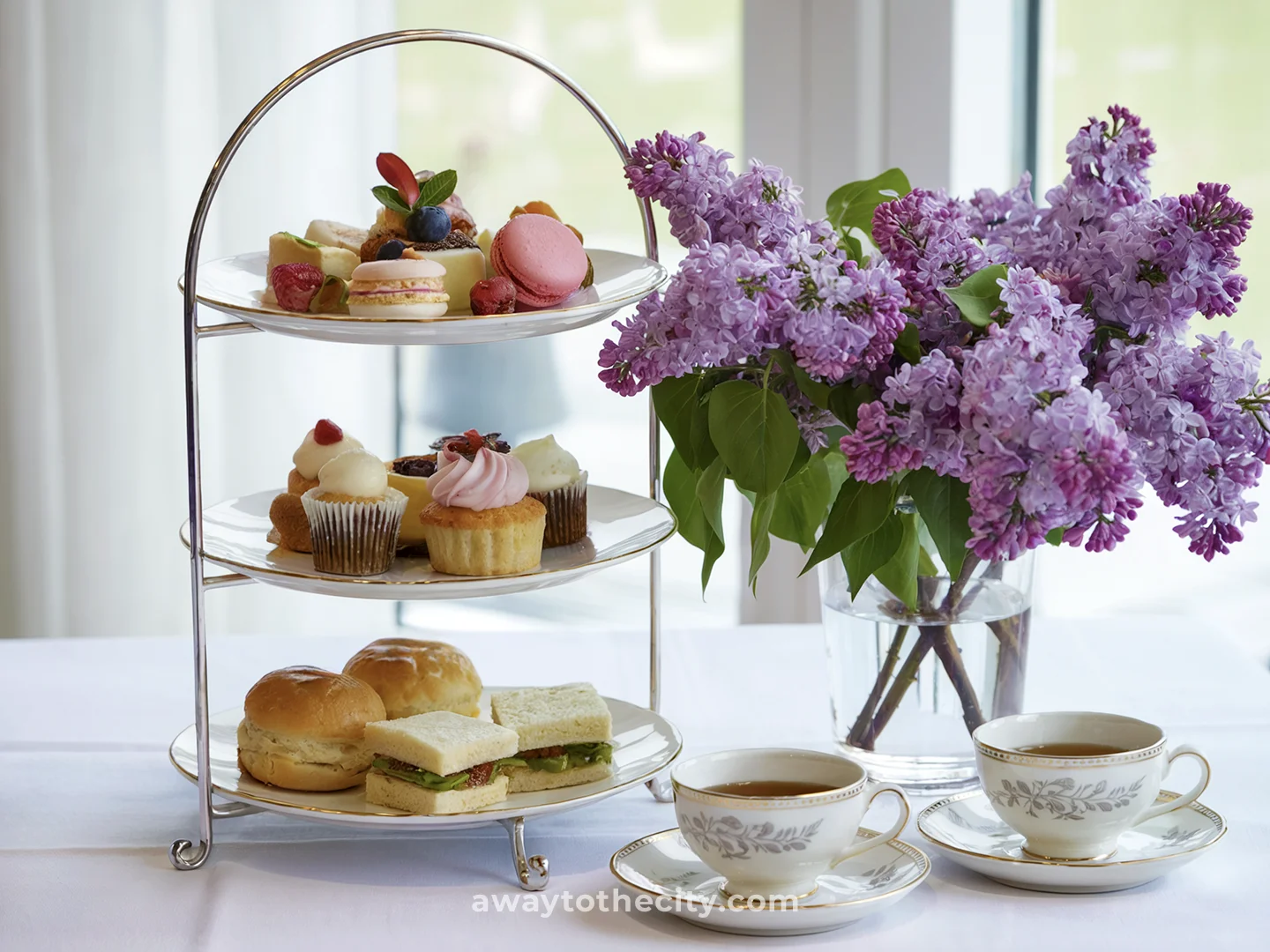 A three-tier stand of savoury and sweet treats for afternoon tea is next to purple flowers and teacups.