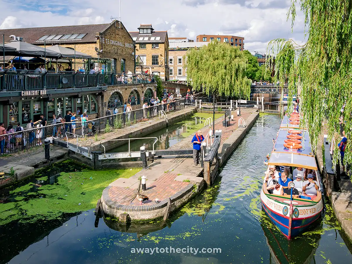 People are gathered around Camden Lock, watching boats on Regent's Canal, with market buildings nearby.