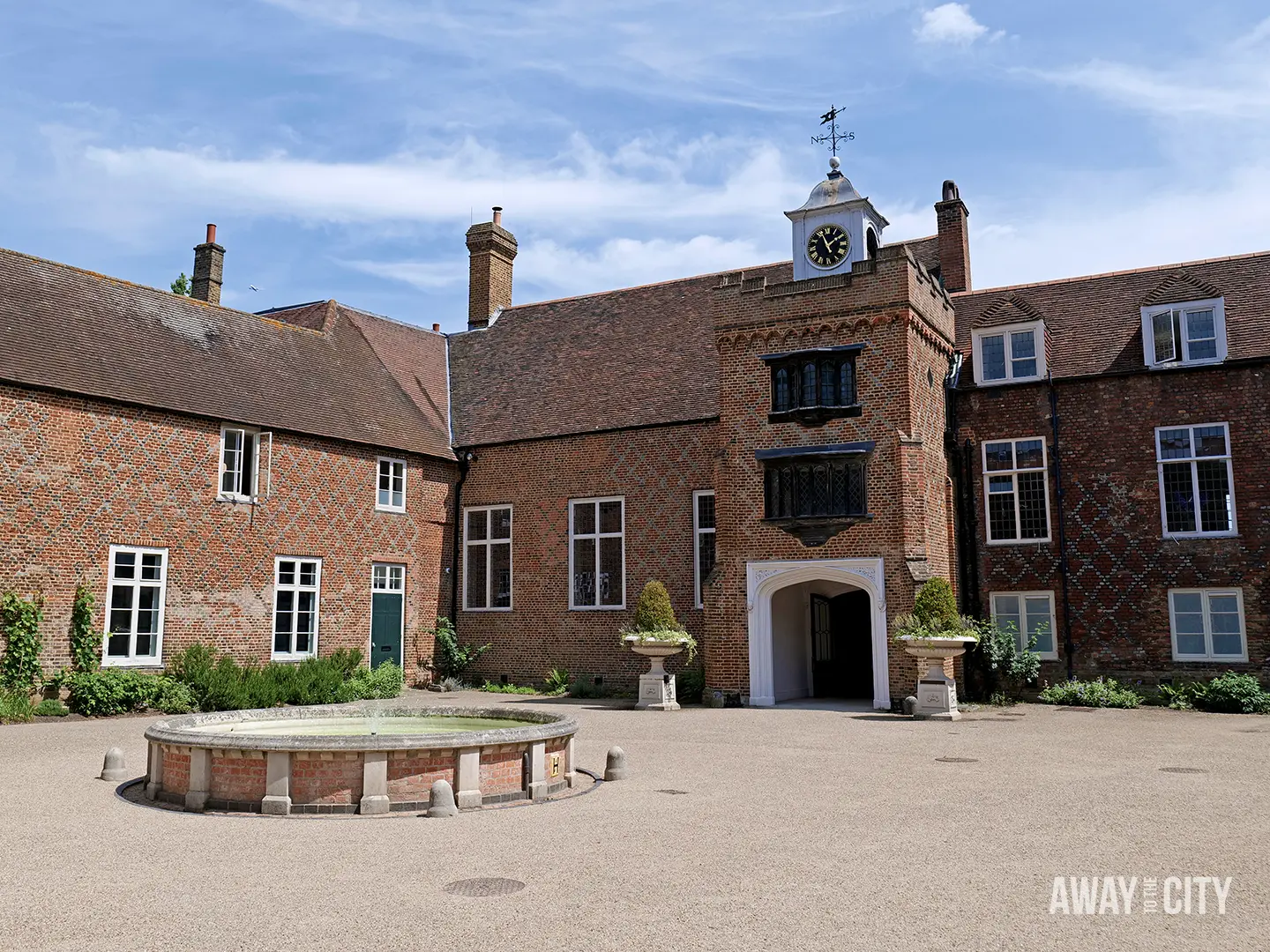 A quaint courtyard at Fulham Palace features a round stone fountain in the middle and a historic red brick building with a clock tower under a blue sky.