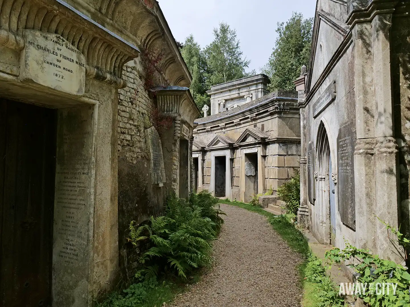 A gravel path winds through the Circle of Lebanon in Highgate Cemetery, flanked by weathered, ornate stone tombs overgrown with green ferns and other plants.