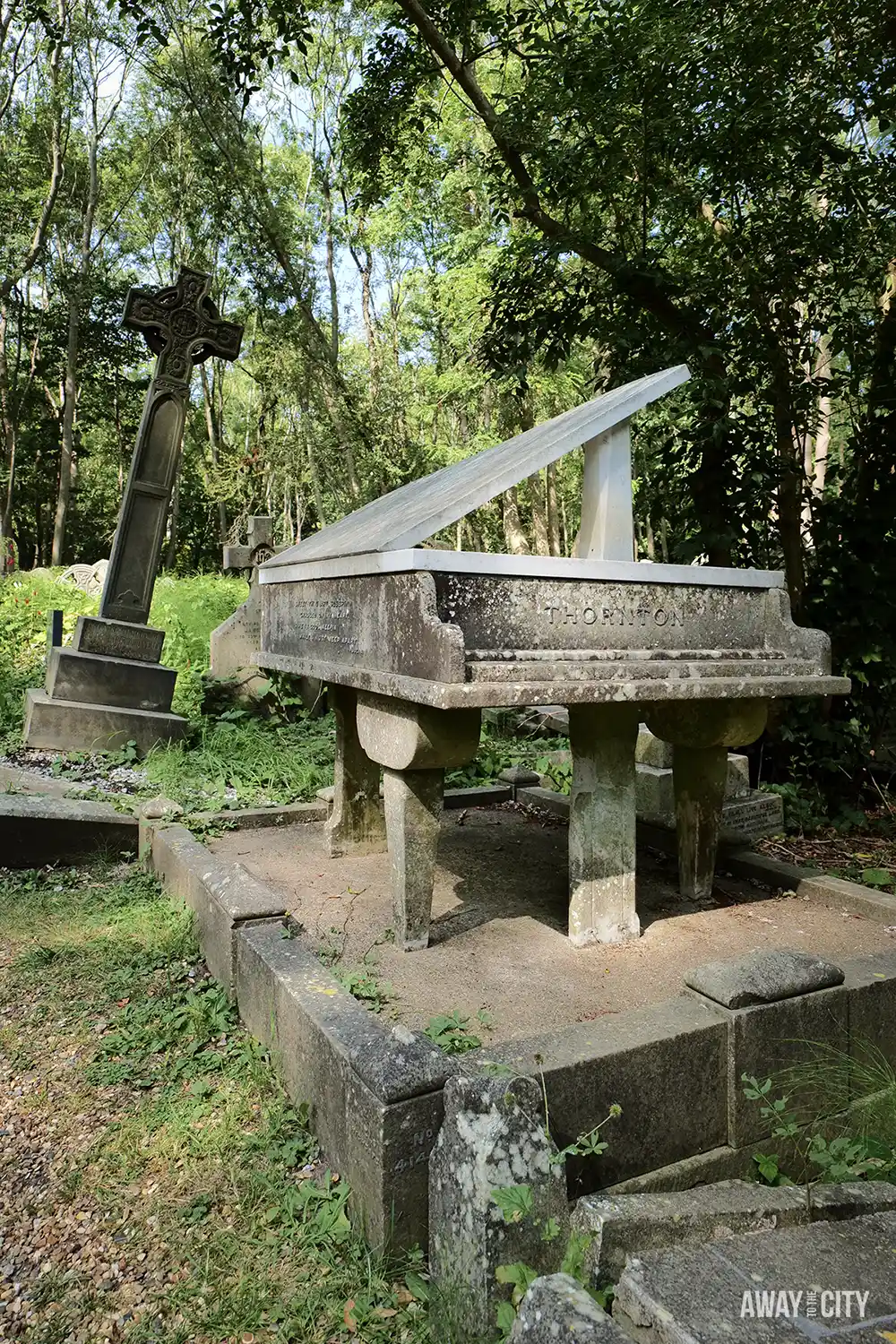 A concrete grand piano-shaped gravestone with "THORNTON" carved into the side sits among other old tombstones and overgrown weeds in a wooded cemetery.