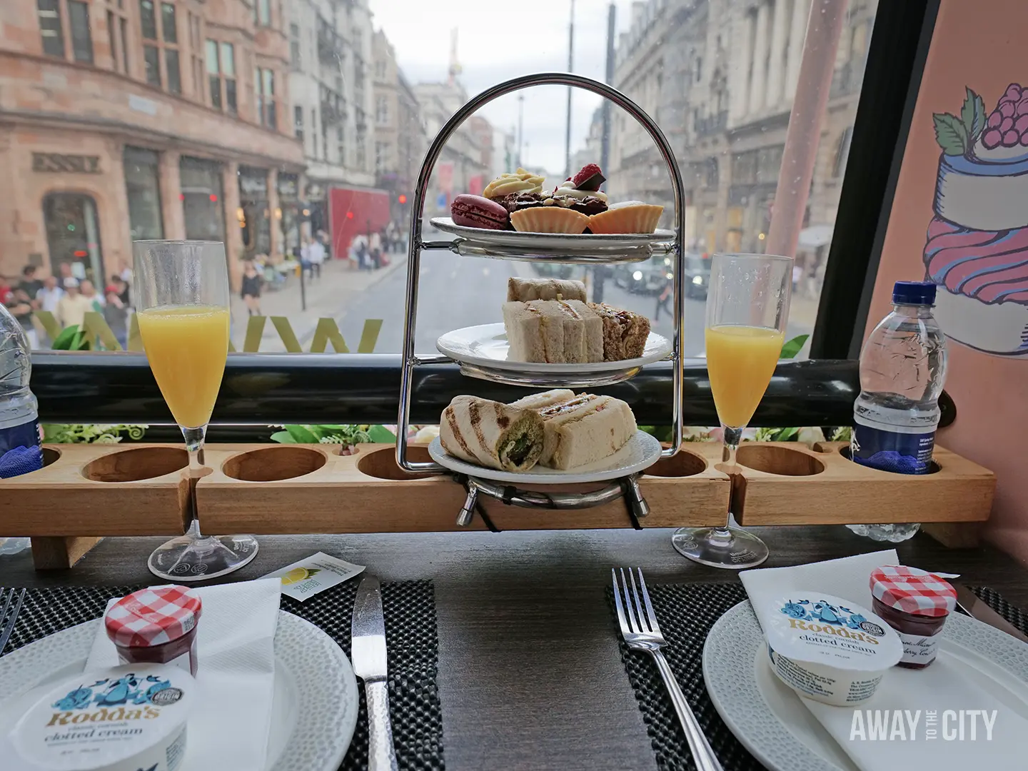 A three-tiered serving platter with assorted pastries, cakes, and sandwiches sits on a table inside a bus, with two glasses of orange juice, a bottle of water, and jars of jam.
