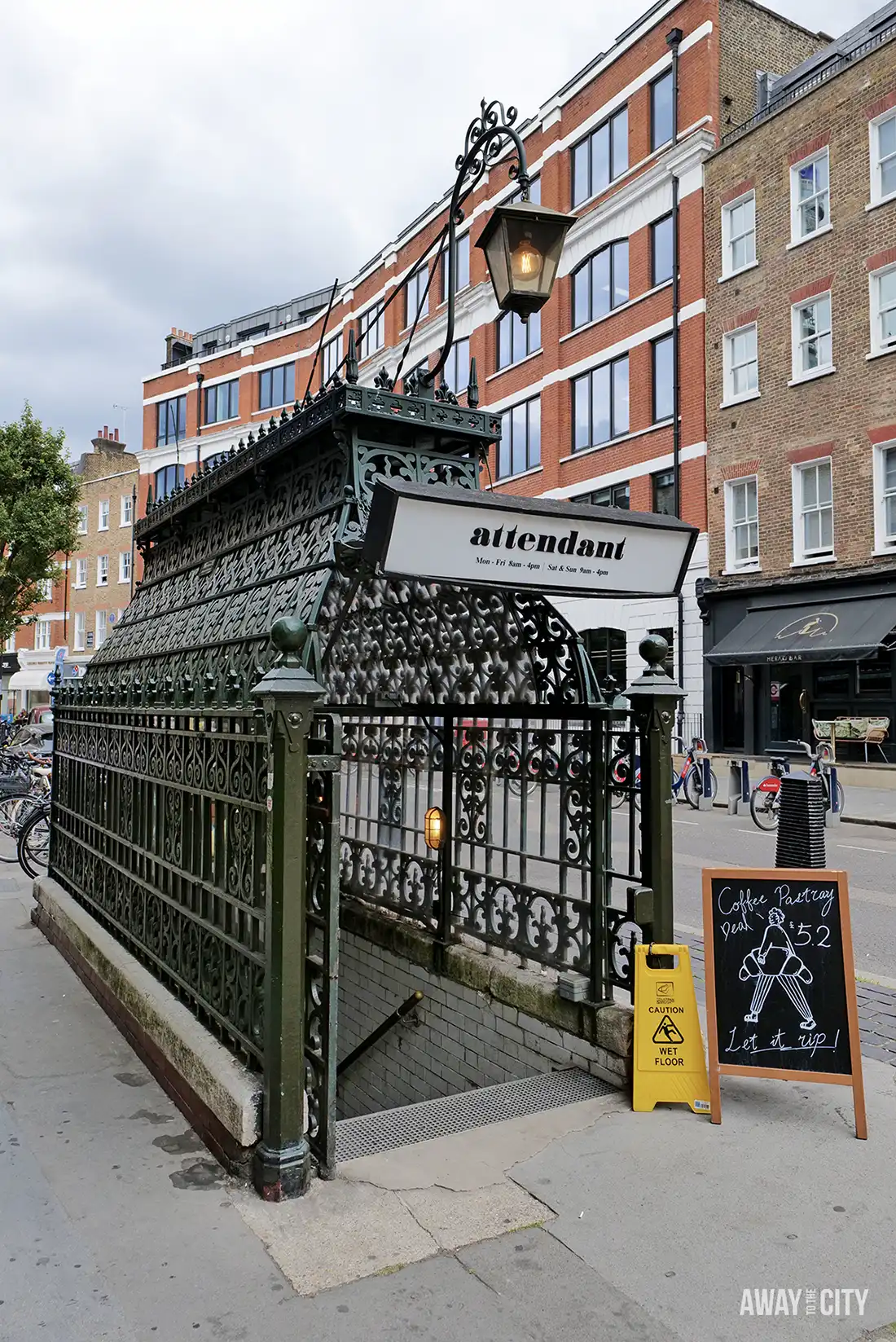 An old public toilet has been converted into a coffee shop, with a black and white sign that says "attendant" over the entrance, leading down a staircase surrounded by a decorative metal fence.