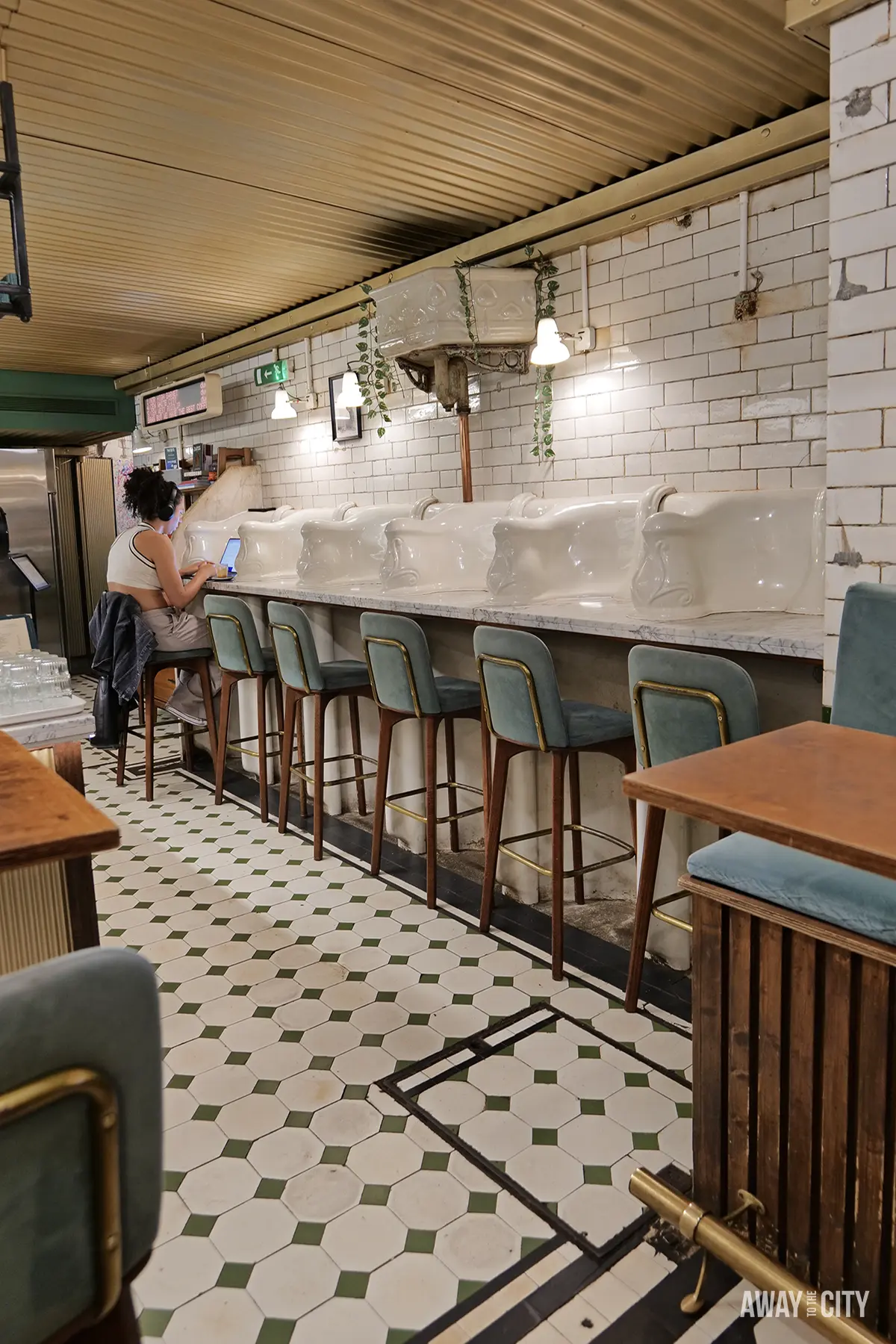A person sits at a counter in a cafe that has white tiled walls and a long counter of repurposed Victorian urinals.
